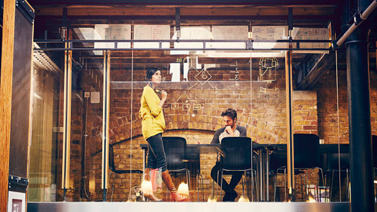 Two creative professionals work on a project, mind mapping ideas onto glass window, in modern office space.